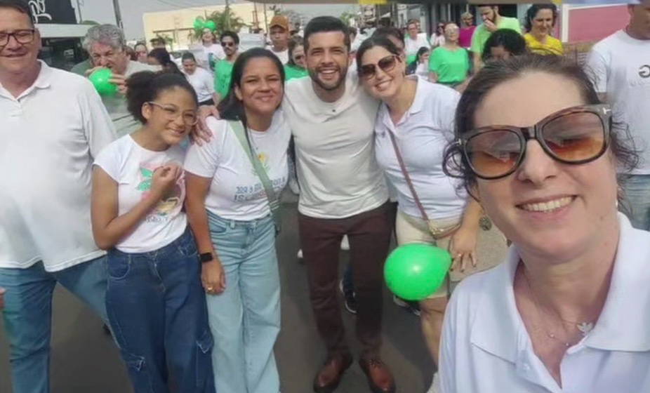 Grupo de pessoas posa em uma rua durante a caminhada da APAE de Tupã. Em primeiro plano, uma mulher de óculos escuros faz uma selfie; participantes e balões verdes aparecem ao fundo.