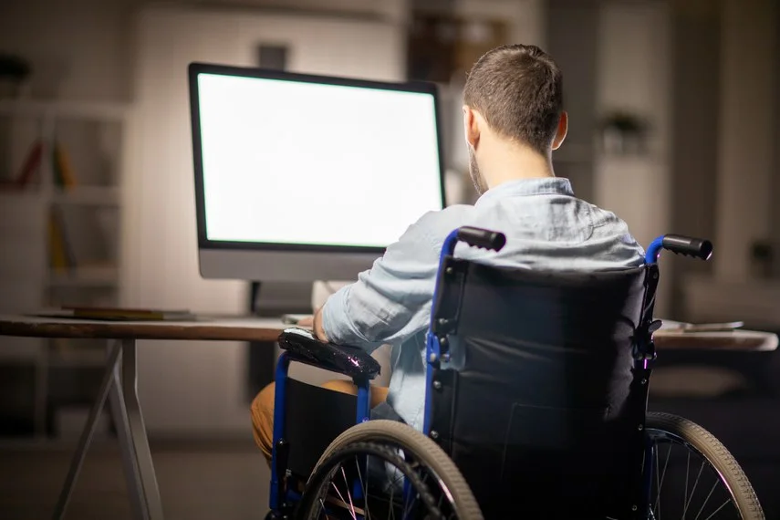 Homem que usa cadeira de rodas está diante de uma mesa, utilizando um computador em ambiente interno.