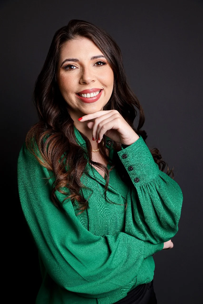 Mulher de cabelos castanhos, longos e ondulados sorri para a câmera. Veste blusa verde, mantém um braço cruzado e a outra mão, com unhas vermelhas, junto ao queixo. Fundo cinza-escuro