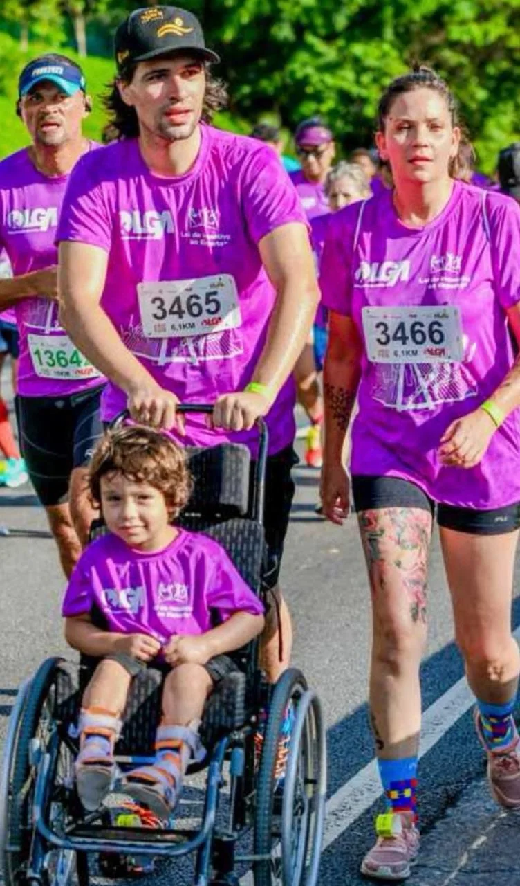 Durante uma corrida de rua, Victor conduz a cadeira de rodas de Jorge enquanto Dayuri corre ao lado deles. Os três usam camisetas roxas do evento. Outros participantes aparecem ao fundo, em uma área arborizada e iluminada pelo sol.