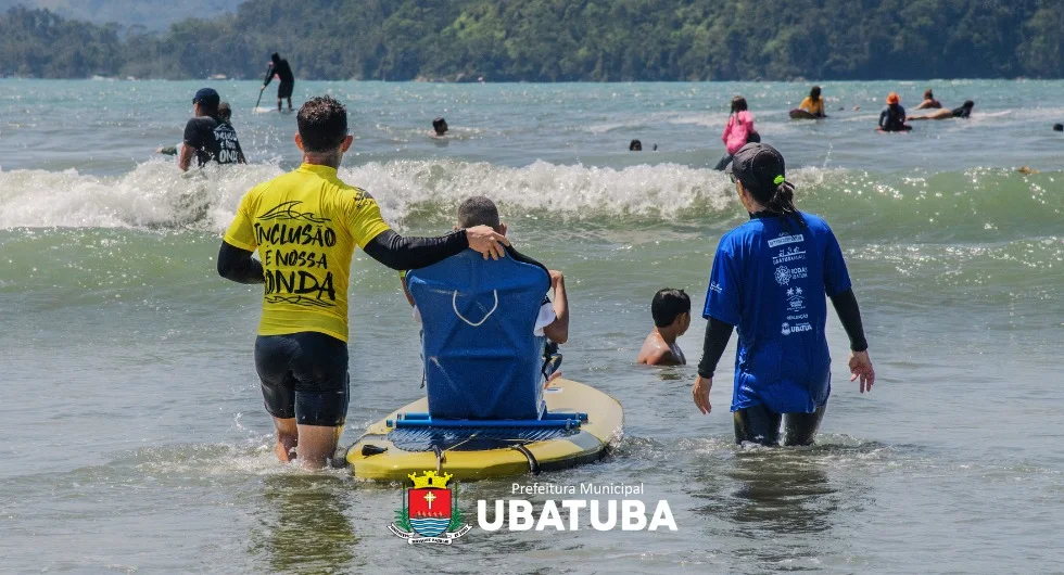 Participante sentado em uma cadeira adaptada sobre uma prancha de surfe, acompanhado por duas pessoas na água do mar.