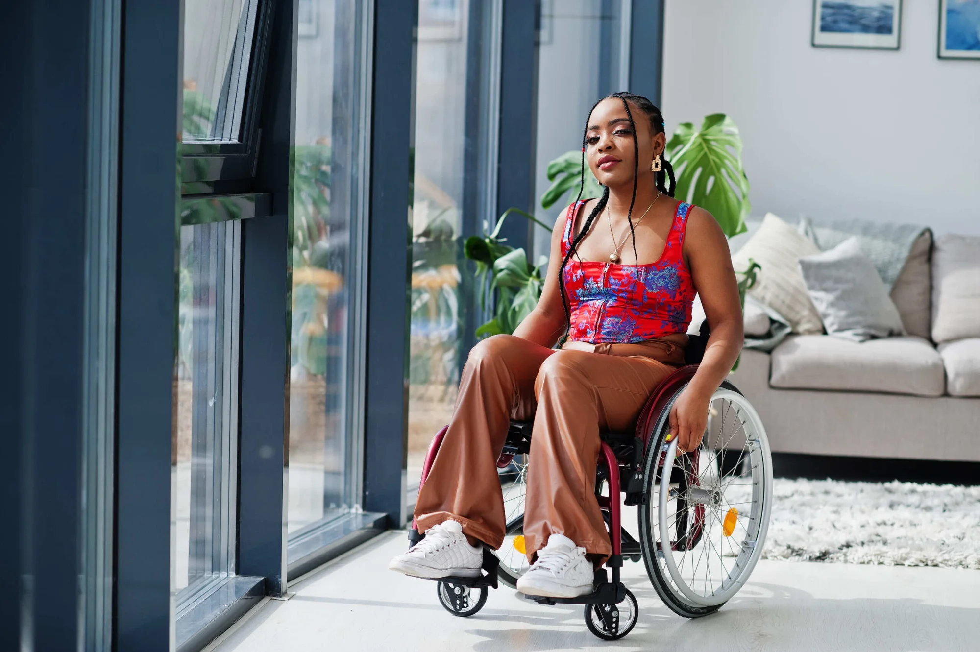 Mulher negra está sentada em uma cadeira de rodas em um ambiente interno bem iluminado. Ela usa blusa vermelha estampada, calça marrom e tênis brancos, além de acessórios como brincos e colar. Seus cabelos estão presos em tranças. À esquerda, há grandes janelas de vidro por onde entra luz natural. A