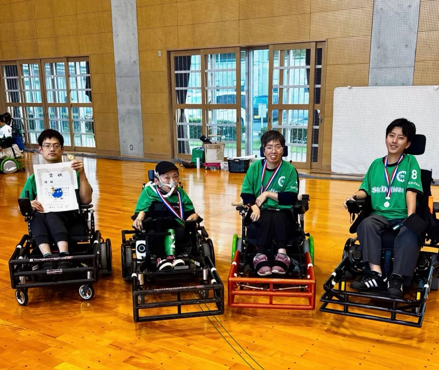 Quatro atletas japoneses em cadeiras de rodas motorizadas, com uniformes verdes, posam em um ginásio após conquista no futebol adaptado. Três exibem medalhas e um segura um certificado.