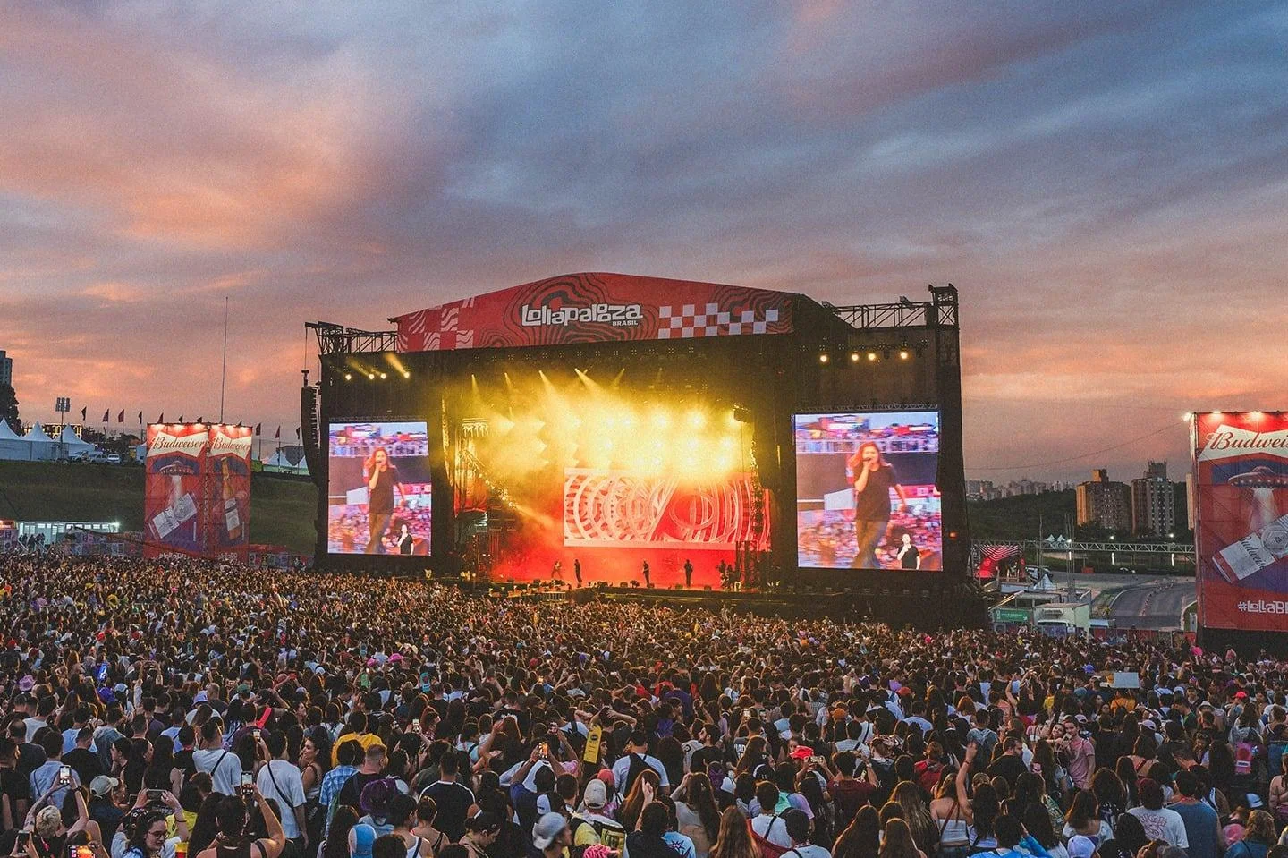 Multidão acompanha show no Lollapalooza Brasil ao entardecer. O palco tem luzes amarelas e vermelhas, dois telões exibem uma cantora e o céu apresenta tons de azul, rosa e laranja.