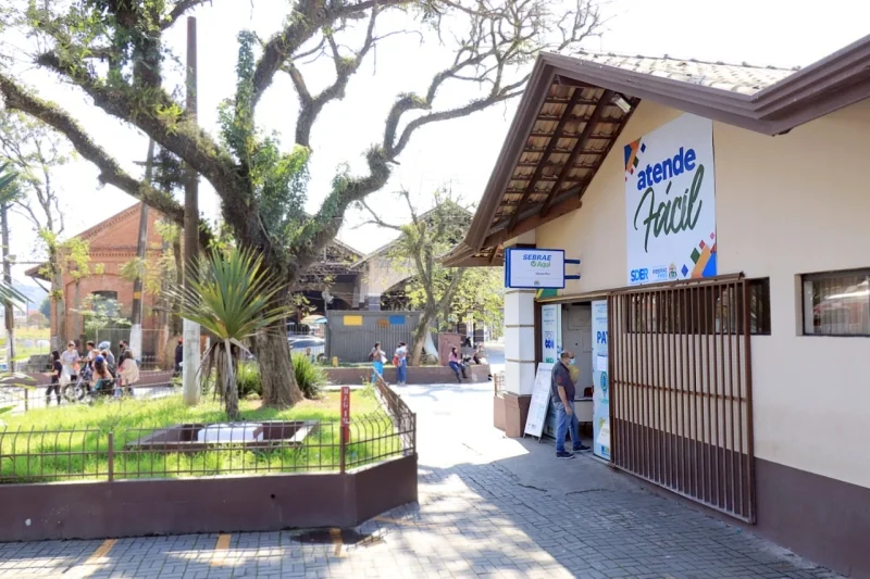 Fachada do Atende Fácil, com placa do Sebrae Aqui, ao lado de uma praça arborizada. Pessoas circulam pela calçada, enquanto uma grande árvore ocupa o centro da imagem.