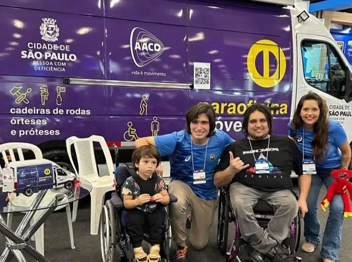 Jorge aparece em sua cadeira de rodas ao lado de Victor e de outras duas pessoas, uma delas também cadeirante. O grupo posa sorridente diante de uma van roxa da Prefeitura de São Paulo e da AACD, utilizada em ações relacionadas a cadeiras de rodas, órteses e próteses.