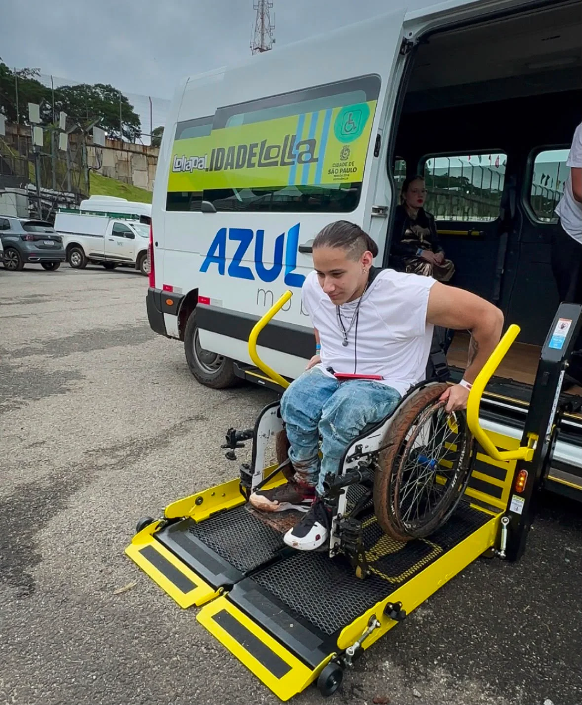 ovem branco, de cabelos castanhos presos para trás, desce em sua cadeira de rodas pela plataforma elevatória de uma van adaptada. Ele veste camiseta branca sem mangas, calça jeans azul e tênis preto e branco. A plataforma é amarela e preta. No veículo branco, aparecem as marcas “Cidade de São Paulo