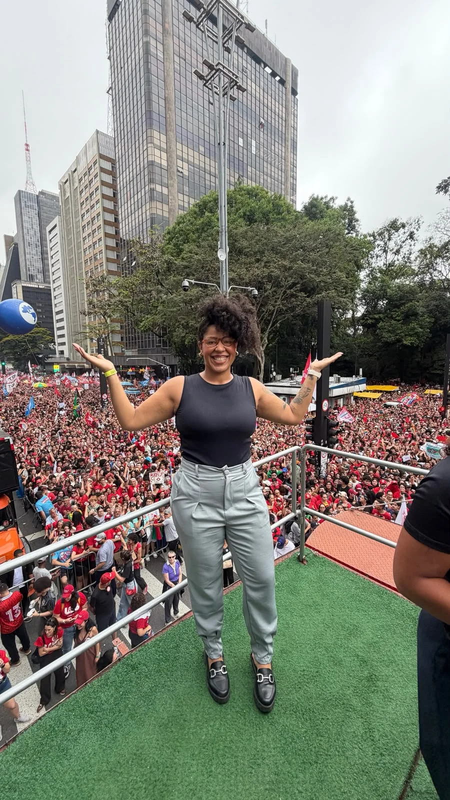 Bia Silva sorri com os braços abertos sobre o carro de som. Ela usa óculos, blusa preta sem mangas e calça cinza. Ao fundo, uma multidão ocupa a Avenida Paulista.
