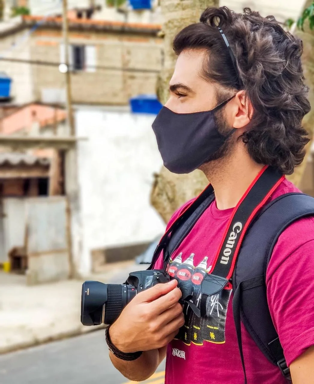 Victor é fotografado de perfil em uma rua, usando camiseta rosa, mochila e máscara preta. Ele segura uma câmera fotográfica profissional junto ao corpo. Ao fundo, casas e construções aparecem desfocadas.