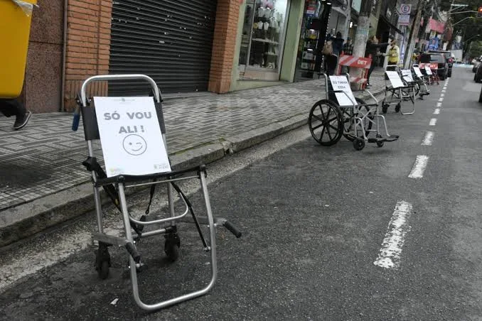 Em uma rua urbana, diversos equipamentos de mobilidade, como cadeiras de rodas e andadores, estão posicionados em sequência junto ao meio-fio. Em cada um deles há uma placa branca com mensagens. No equipamento mais próximo da câmera, a placa traz a frase “SÓ VOU ALI!”, acompanhada de um rosto sorrid