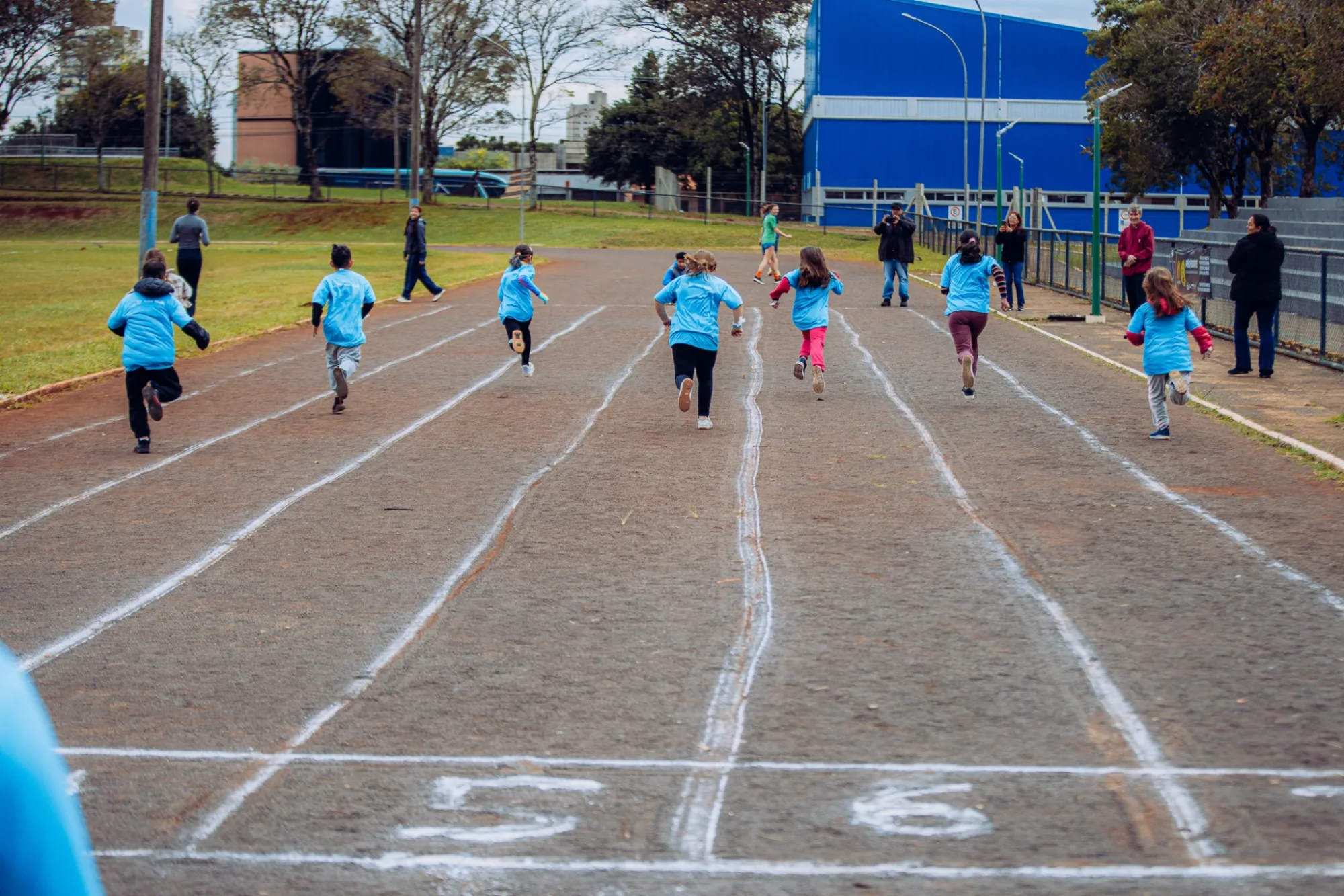Crianças participam de uma corrida em uma pista de atletismo ao ar livre. Elas correm de costas para a câmera, distribuídas pelas raias e usando camisetas azuis. Algumas vestem calças ou bermudas em diferentes cores. À direita e ao fundo da pista, adultos acompanham a atividade, observando, fotograf