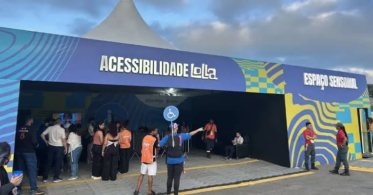 Fotografia da entrada da Central de Acessibilidade do Lollapalooza Brasil. A estrutura possui fachada azul e roxa, com as inscrições “ACESSIBILIDADE Lolla” e “ESPAÇO SENSORIAL” em letras brancas. No centro, aparece o símbolo internacional de acesso, representado por uma cadeira de rodas.