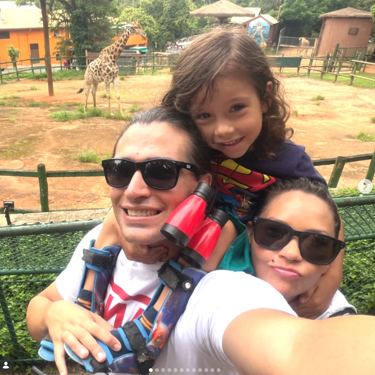 Fotografia ao ar livre mostra uma mulher sorrindo ao lado de uma criança, que está sentada à sua frente em uma cadeira de rodas com estrutura frontal e guidão. A mulher tem cabelos longos e escuros, usa roupa em tons de azul e segura uma lata de bebida. A criança tem cabelos castanhos, veste moletom