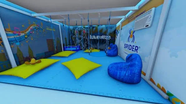 Fotografia de uma sala sensorial do Lollapalooza Brasil, com piso azul acolchoado, tapetes amarelos espalhados pelo chão e grandes pufes azuis. O ambiente possui paredes decoradas com ilustrações coloridas, equipamentos sensoriais suspensos e iluminação azul suave.