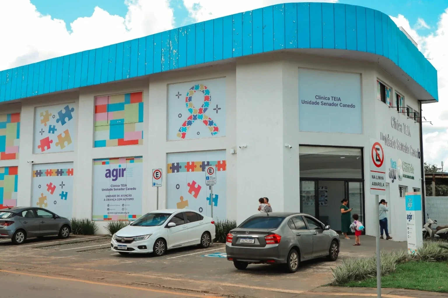 Fachada branca e azul da Clínica TEIA de Senador Canedo, com painéis coloridos, carros estacionados e pessoas próximas à entrada.