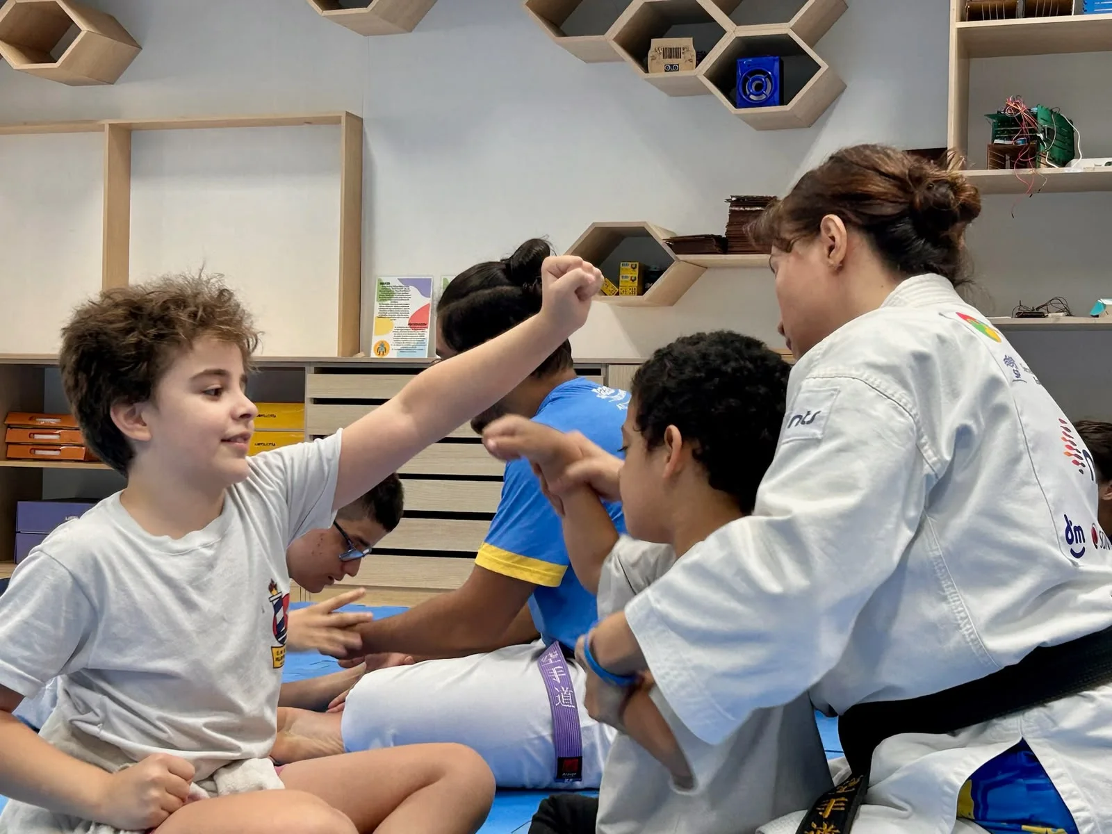 Alunos praticam movimentos de karatê sentados em tatame azul, acompanhados por uma instrutora de quimono branco.