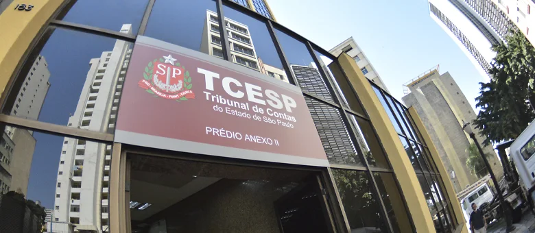 Fachada de um prédio do Tribunal de Contas do Estado de São Paulo (TCE-SP), vista de baixo para cima. Em destaque, uma placa em tons de vermelho e branco identifica o órgão e traz a inscrição “TCE-SP – Tribunal de Contas do Estado de São Paulo – Prédio Anexo II”. A fachada possui grandes painéis de