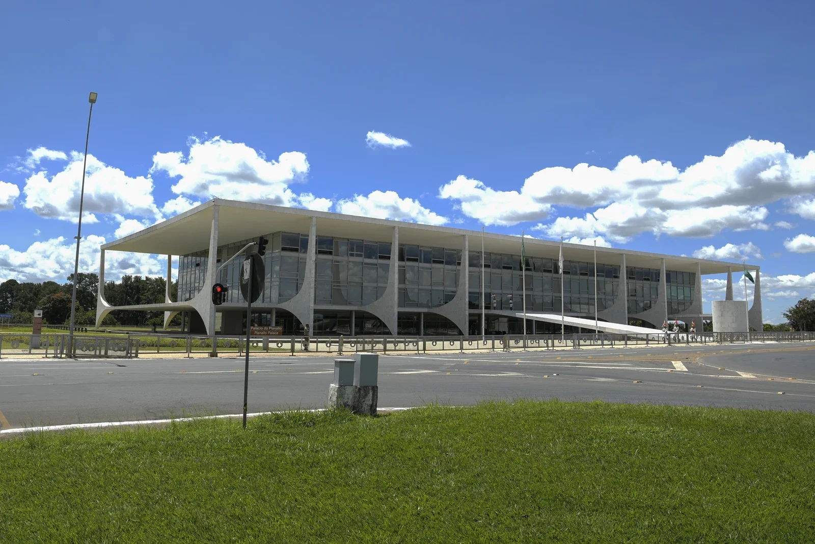 Fachada do Palácio do Planalto, em Brasília, com pilares brancos, paredes de vidro e rampa de acesso, vista do outro lado da rua sob céu azul com nuvens.