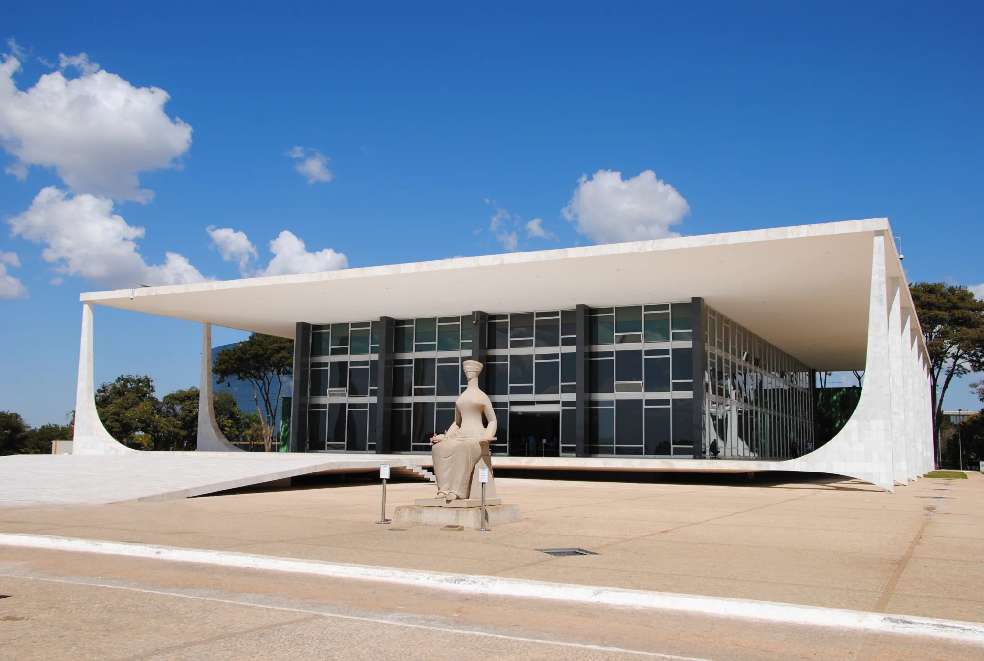 Fotografia da fachada do Supremo Tribunal Federal (STF), em Brasília, em um dia ensolarado. O edifício tem grandes paredes de vidro, uma ampla cobertura horizontal e colunas brancas com curvas na base. Em primeiro plano, ao centro, está a escultura da Justiça, representada por uma mulher sentada, co