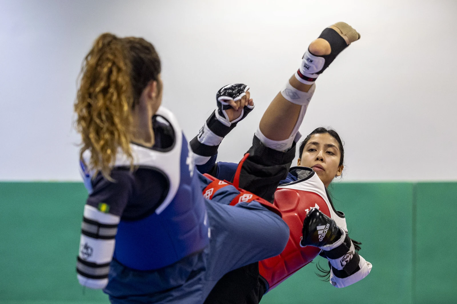 Duas atletas treinam taekwondo com protetores azul e vermelho; uma delas mantém a perna elevada em um chute.