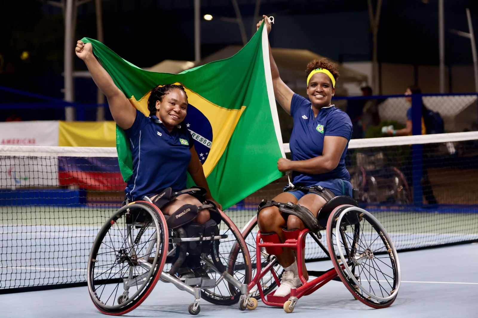 Vitória Miranda e Meirycoll Duval, sentadas em cadeiras de rodas, erguem a bandeira do Brasil em uma quadra de tênis.