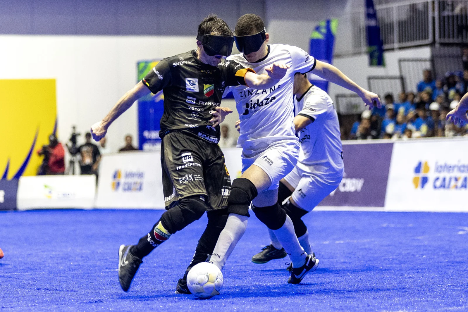 Dois jogadores vendados, com uniformes preto e branco, disputam a bola na quadra de futebol de cegos.