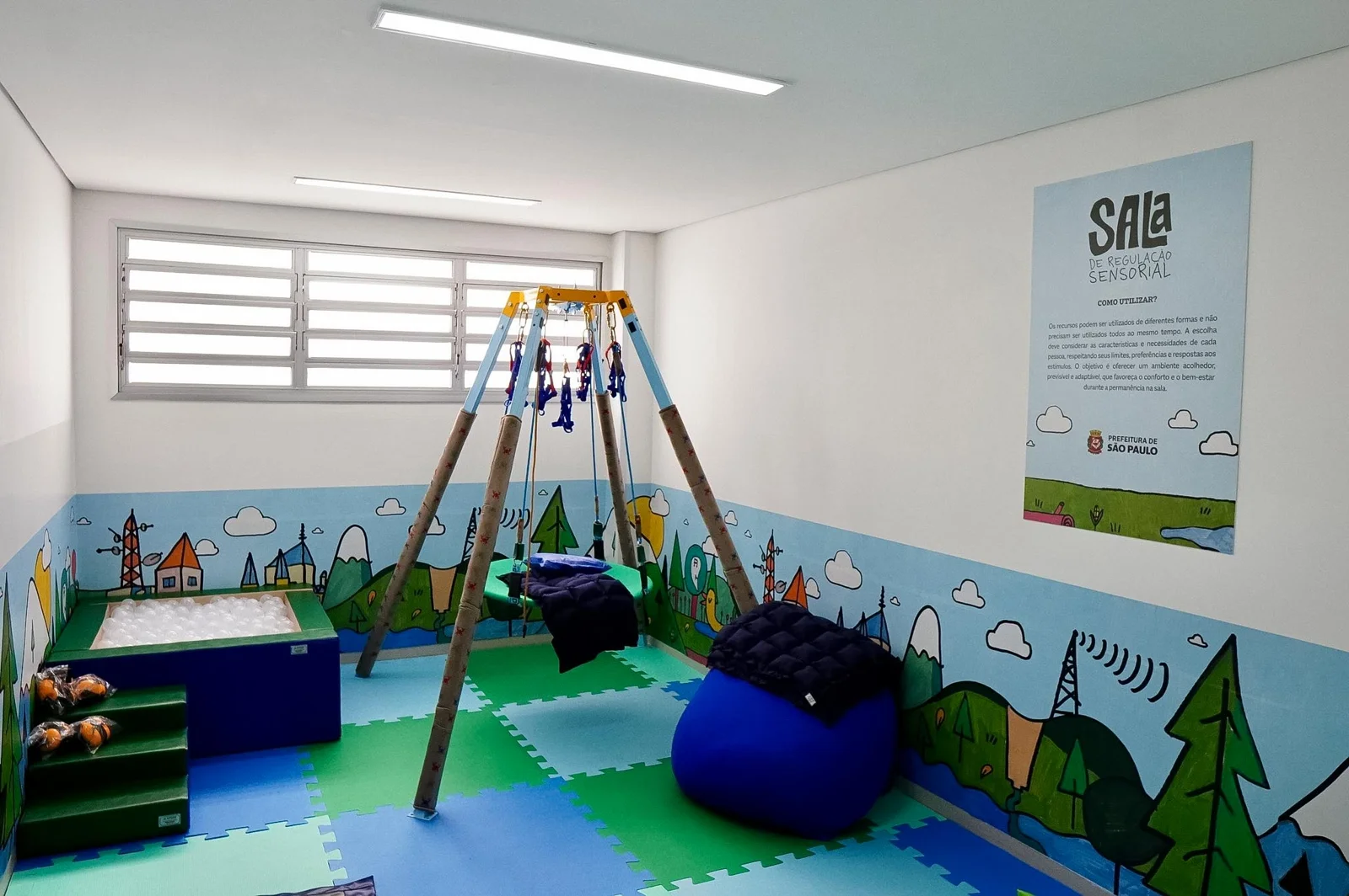 Sala sensorial da UBS Jardim Helena, com piso de placas azuis e verdes, balanço, pufe azul e desenhos de paisagens nas paredes.