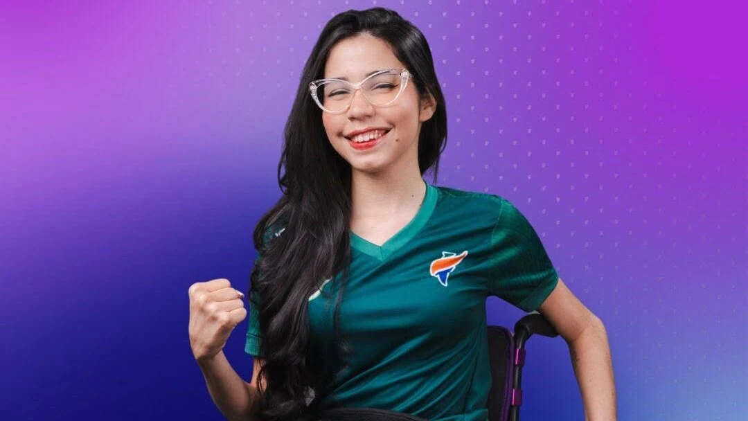 Laíssa Guerreira sorri, com óculos transparentes e camiseta verde, sentada em cadeira de rodas, com o punho direito erguido diante de fundo roxo.