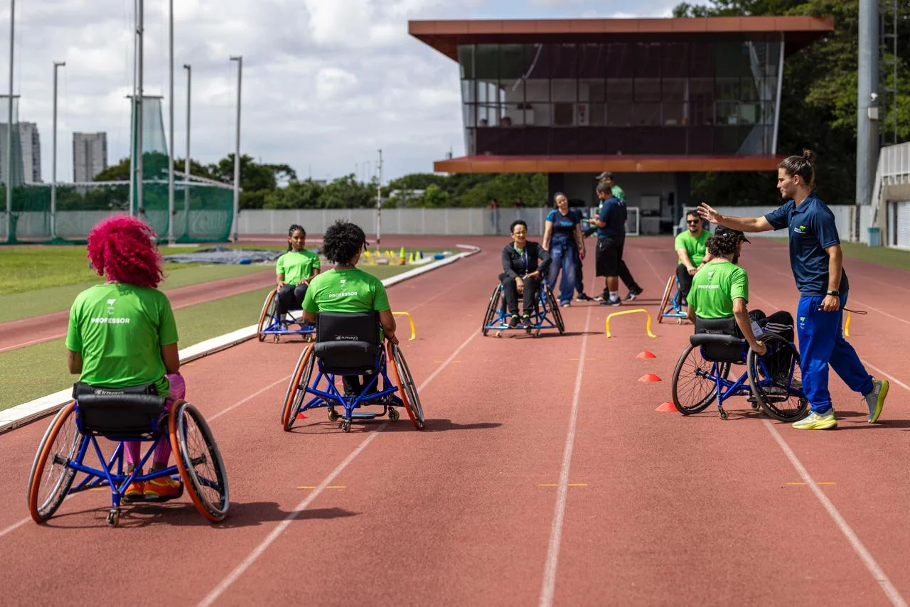 Professores participam de atividade com cadeiras de rodas esportivas em uma pista de atletismo, sob orientação de instrutores.