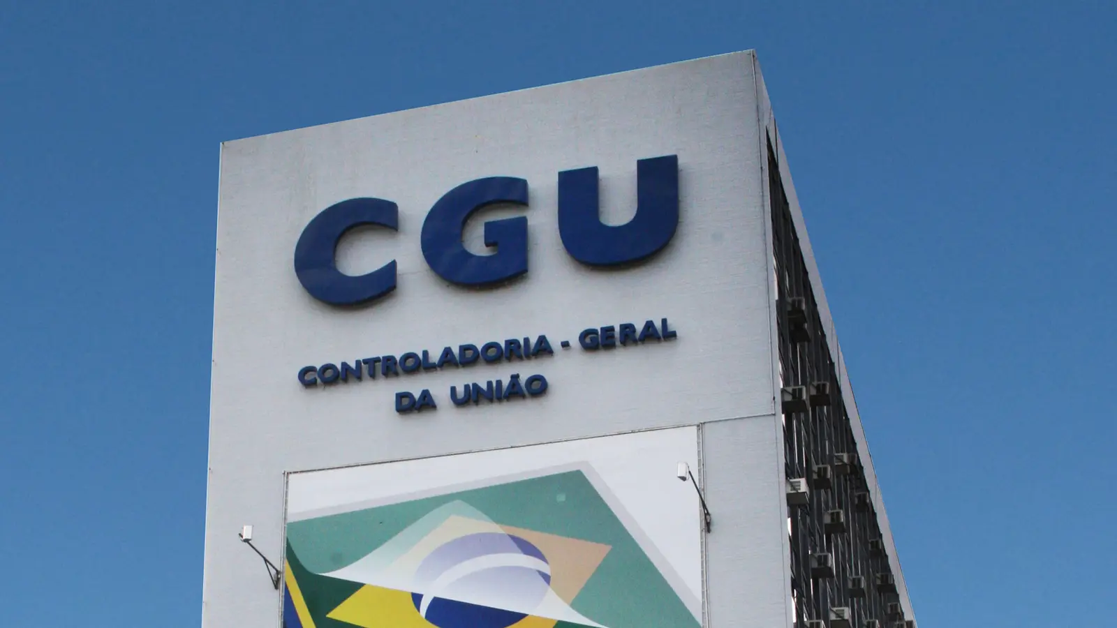 Detalhe da fachada branca da Controladoria-Geral da União, com a sigla CGU e o nome do órgão em letras azuis, sob céu azul.
