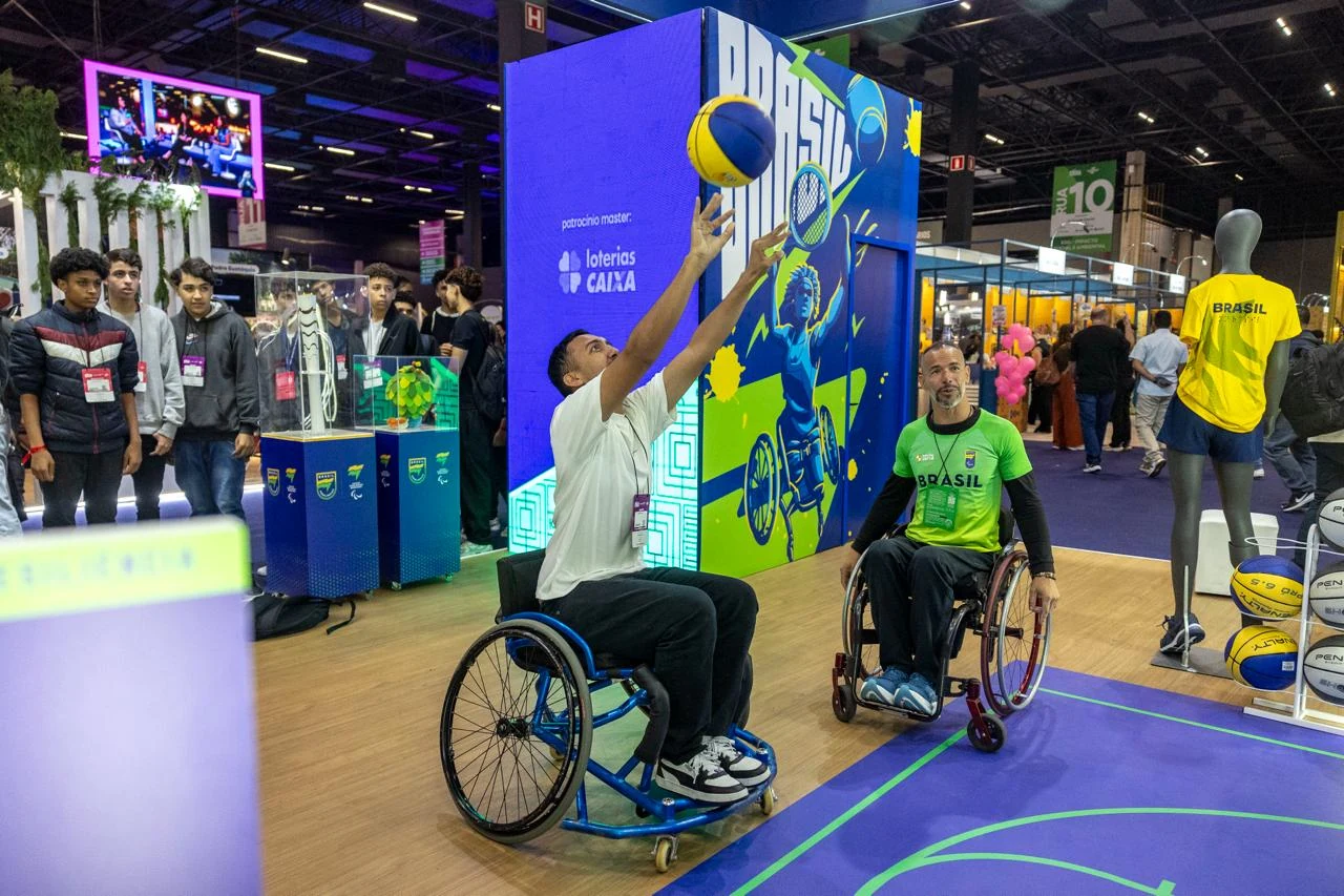 Visitante em cadeira de rodas esportiva arremessa uma bola de basquete no estande Brasil Paralímpico; ao lado, outro participante acompanha a atividade.