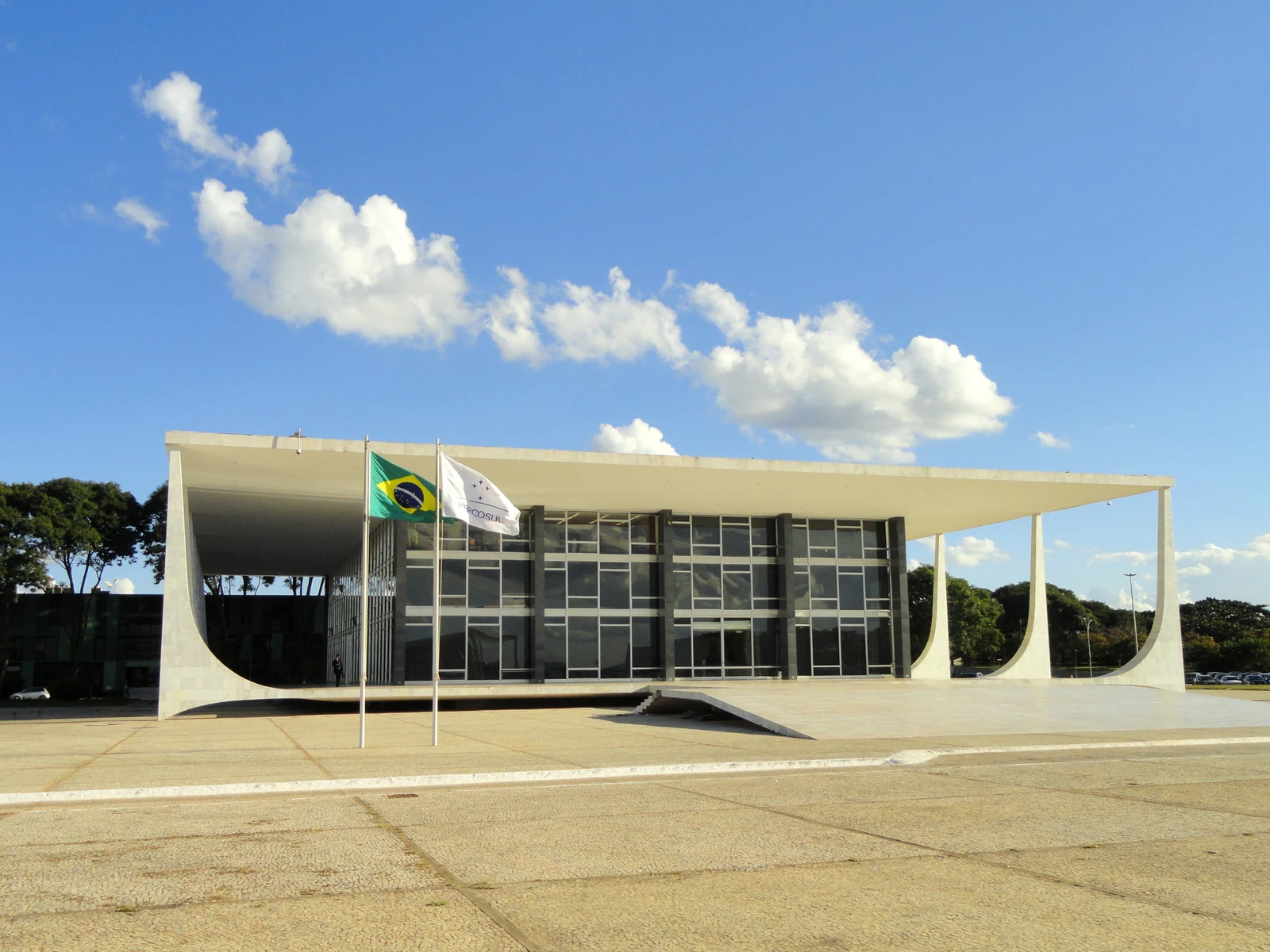 Fachada do Supremo Tribunal Federal, em Brasília, com colunas brancas, paredes de vidro e bandeiras diante do edifício, sob céu azul.