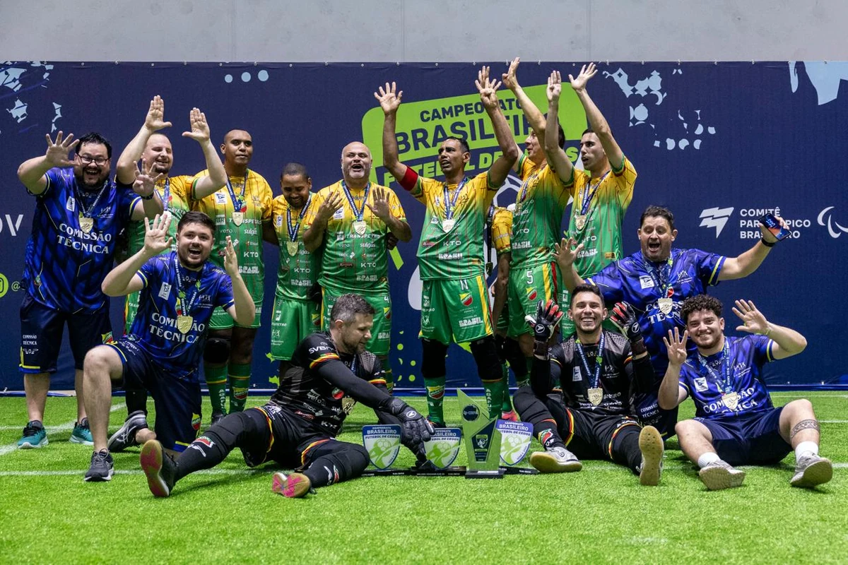 Atletas e comissão técnica da AGAFUC comemoram com medalhas e troféus no gramado do CT Paralímpico. Jogadores de verde e amarelo levantam as mãos, ao lado de integrantes da equipe de azul.