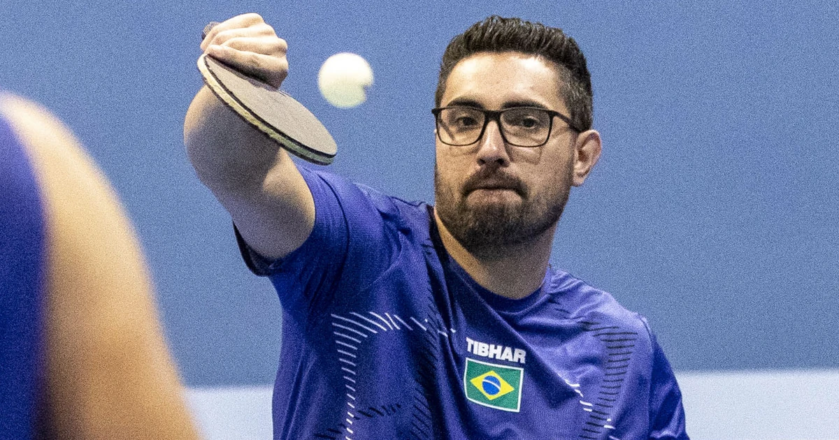 Fábio Souza da Silva, de óculos e camiseta azul com a bandeira do Brasil, estende a raquete em direção a uma bola de tênis de mesa. Foto de arquivo.