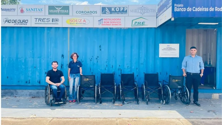 Duas pessoas em pé e um homem em cadeira de rodas posam ao lado de cadeiras de rodas diante da fachada azul do banco de cadeiras de rodas do Rotary Club de Juína.