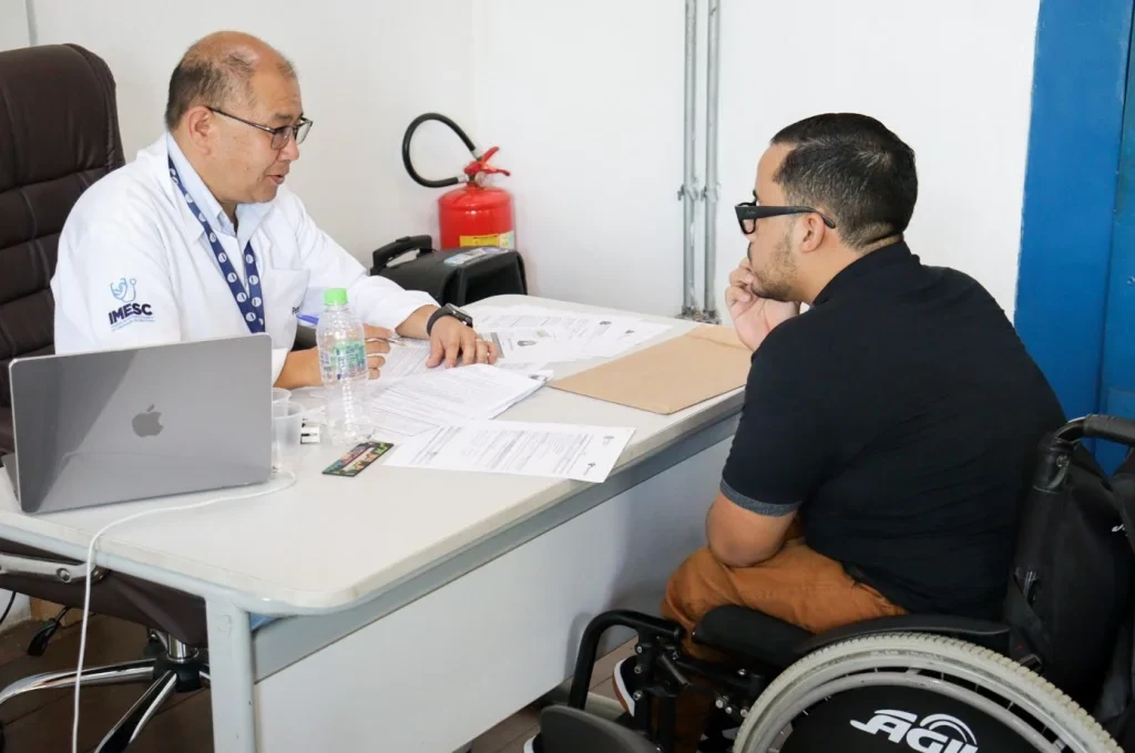 Profissional de jaleco do Imesc conversa com um homem em cadeira de rodas diante de uma mesa com documentos e computador.