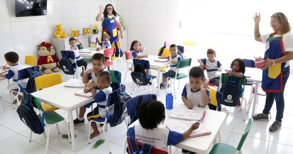 Crianças com uniforme azul e branco desenham em mesas de uma sala de creche, acompanhadas por duas educadoras. Foto de arquivo de Crato, Ceará.