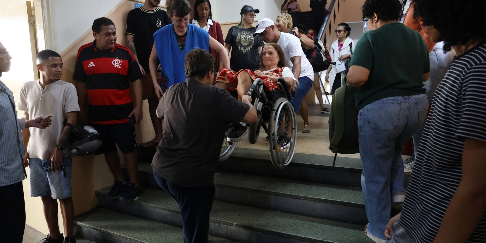 Maria Lidia Gomes de Mattos, em cadeira de rodas, recebe auxílio de pessoas em uma escada no local de votação.