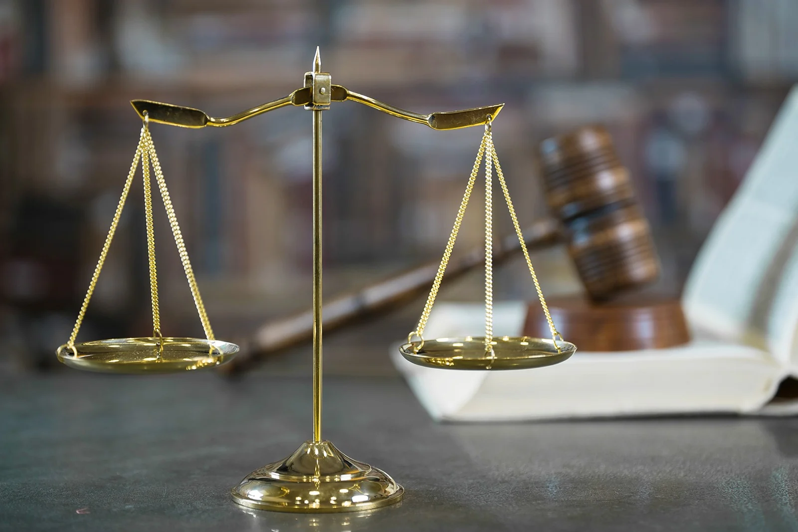 Balança dourada da Justiça em primeiro plano, com um martelo e um livro desfocados ao fundo.