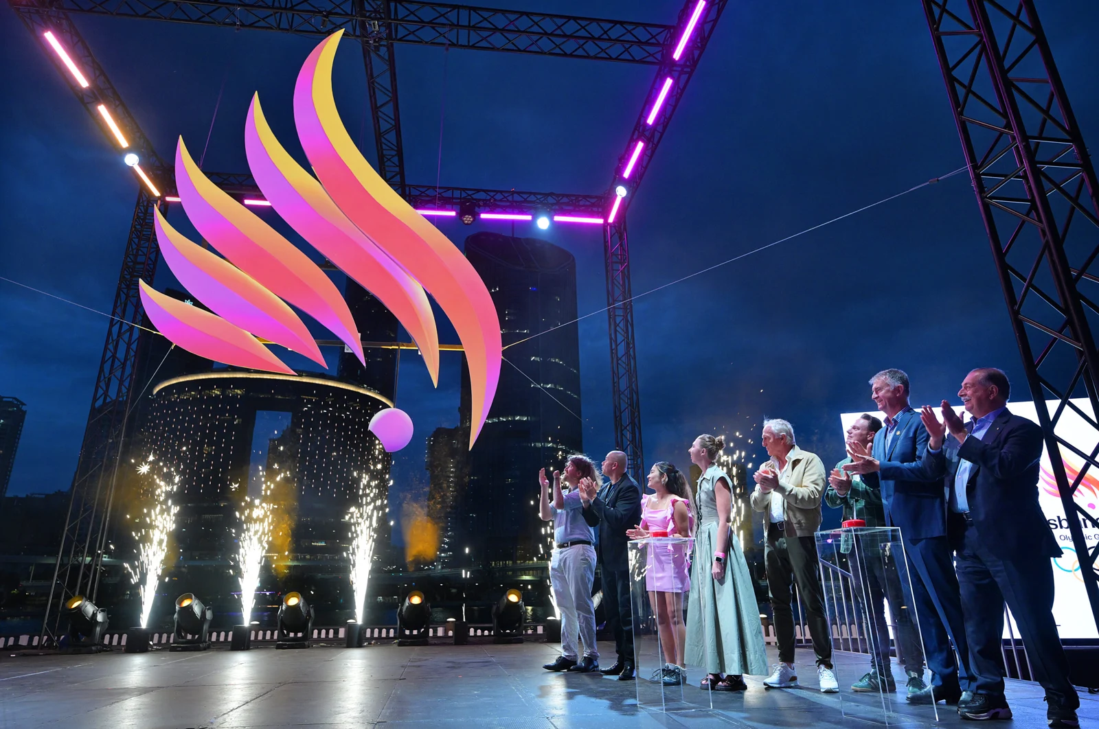 Convidados aplaudem o lançamento do emblema iluminado dos Jogos de Brisbane 2032 em um palco.