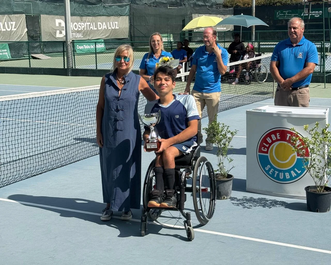 João Takaki, em sua cadeira de rodas, segura um troféu ao lado de uma mulher na quadra de tênis.