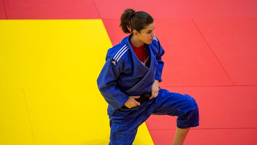 Judoca com quimono azul e faixa preta sobre o tatame vermelho e amarelo.