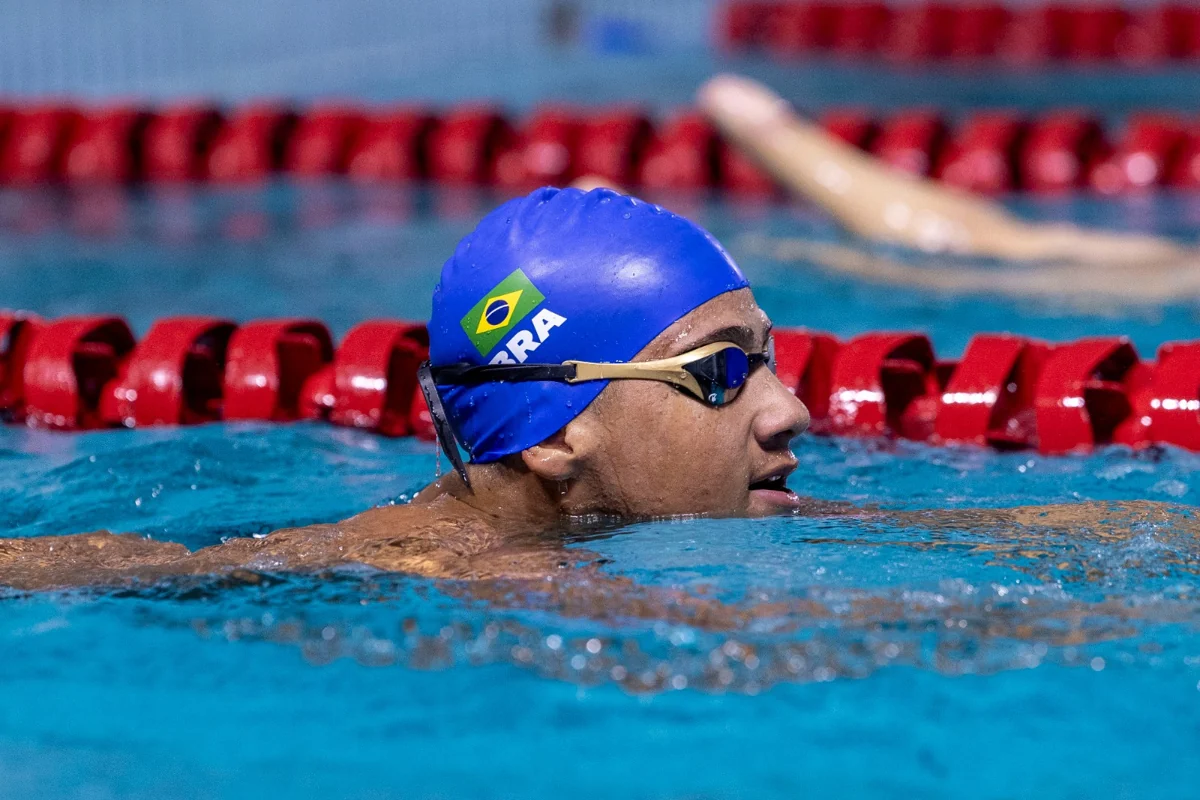 Luiz Fernando Antonio Rodrigues na piscina, de touca azul com bandeira do Brasil e óculos de natação, junto a uma raia vermelha.