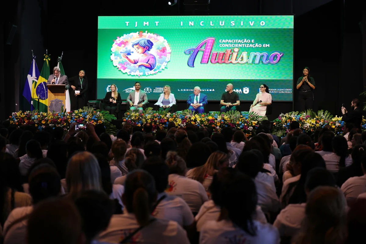 Público sentado diante de um palco com autoridades, intérpretes de Libras e telão do evento TJMT Inclusivo.