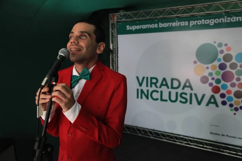 Homem de paletó vermelho e gravata borboleta verde segura um microfone diante de um painel da Virada Inclusiva.