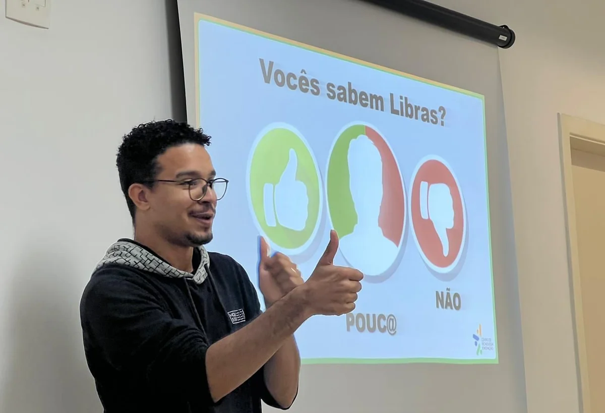 Homem de óculos faz gestos com as mãos diante de uma projeção com a pergunta “Vocês sabem Libras?”.