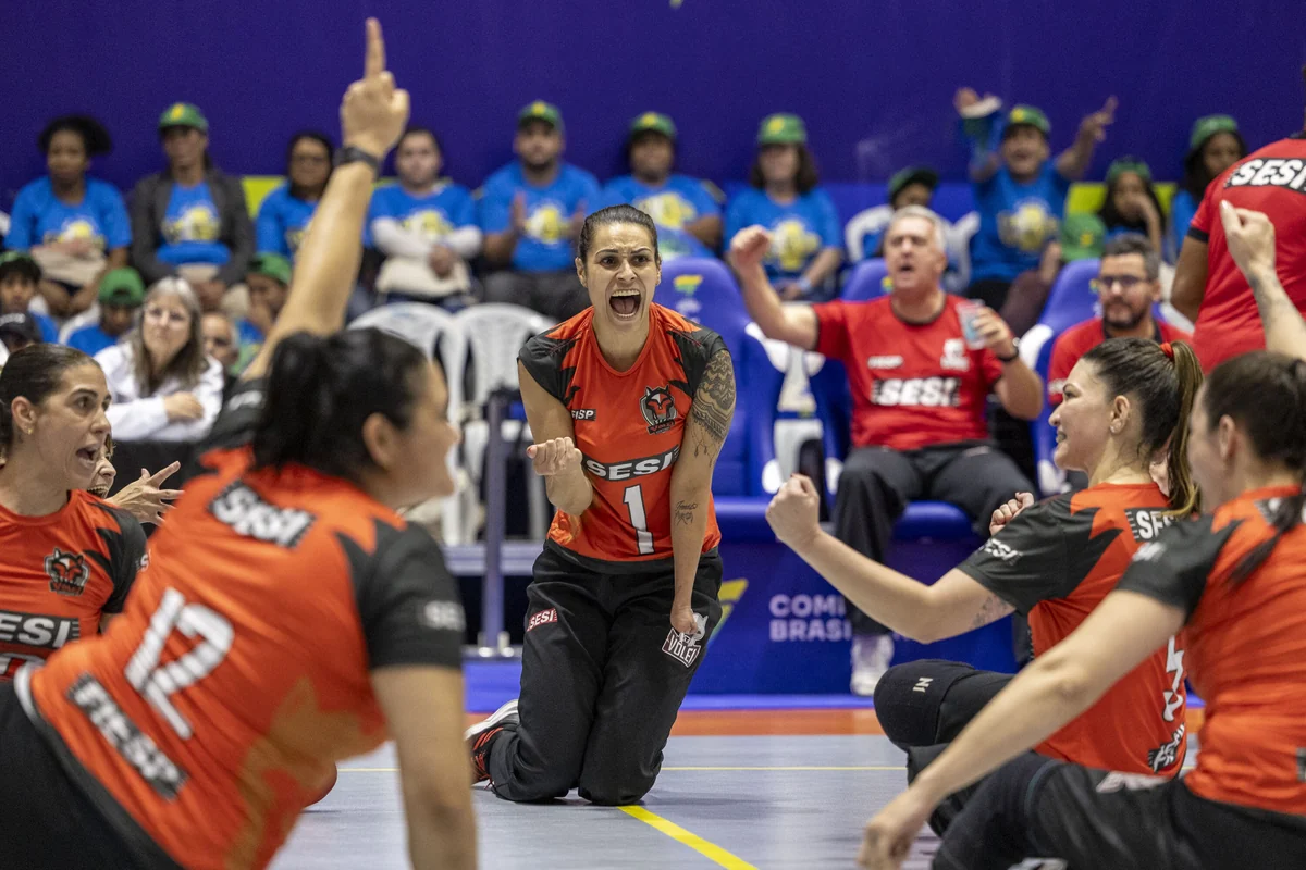 Jogadoras do SESI-SP, com uniformes vermelhos e pretos, comemoram na quadra de vôlei sentado diante da torcida.