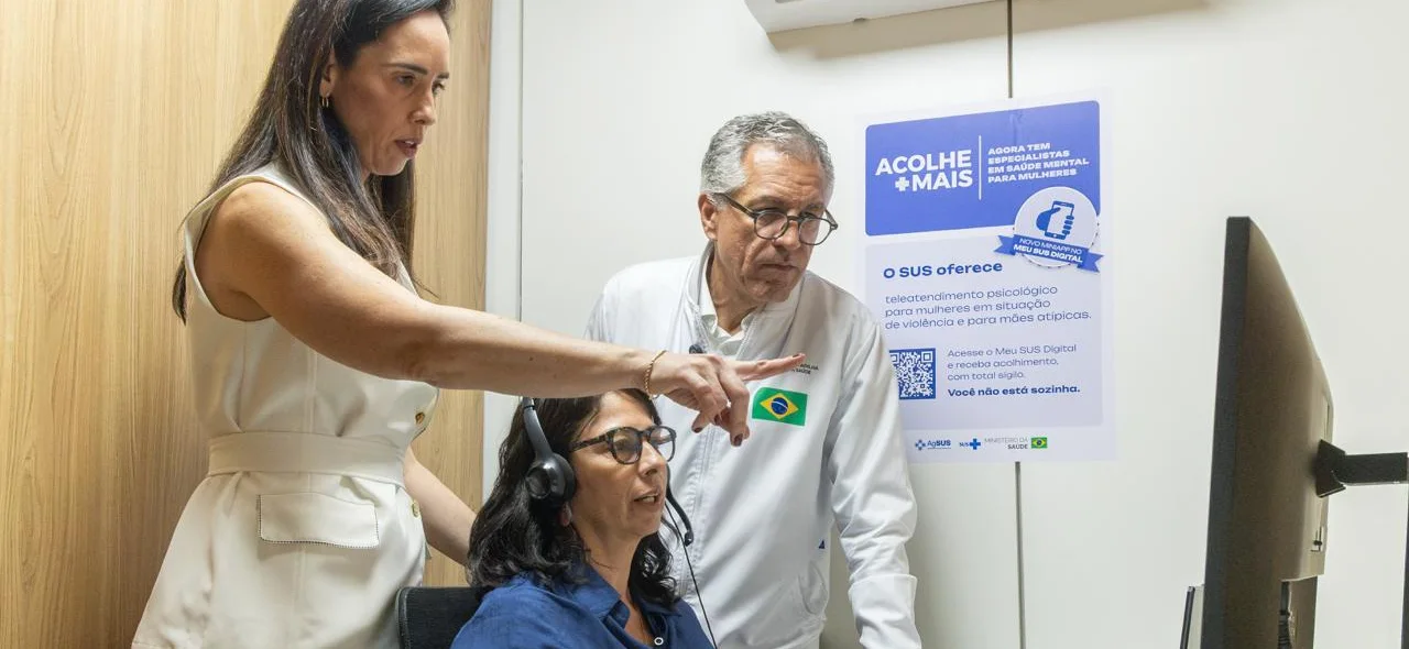 Três pessoas diante de um computador, ao lado de um cartaz do Acolhe Mais.