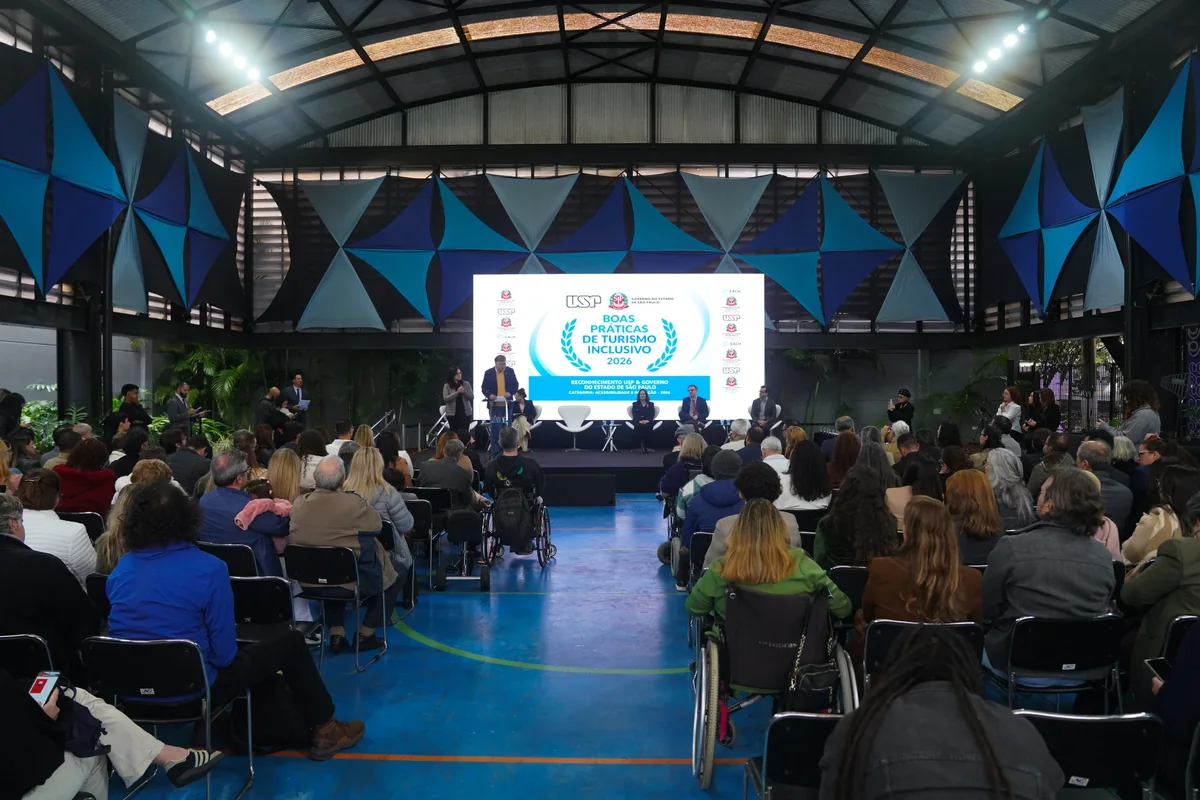 Público sentado, incluindo pessoas em cadeiras de rodas, acompanha uma cerimônia diante de palco e painel de boas práticas de turismo inclusivo.