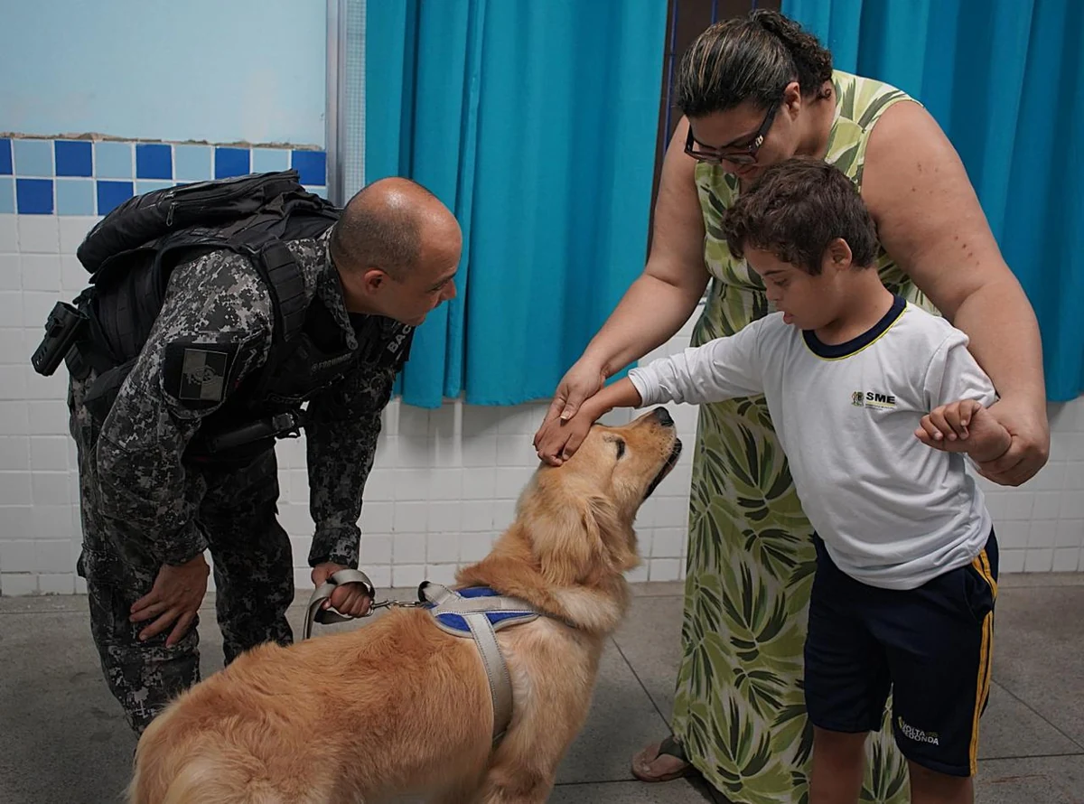 Menino toca um cão com apoio de uma mulher, ao lado de um policial que segura a guia.